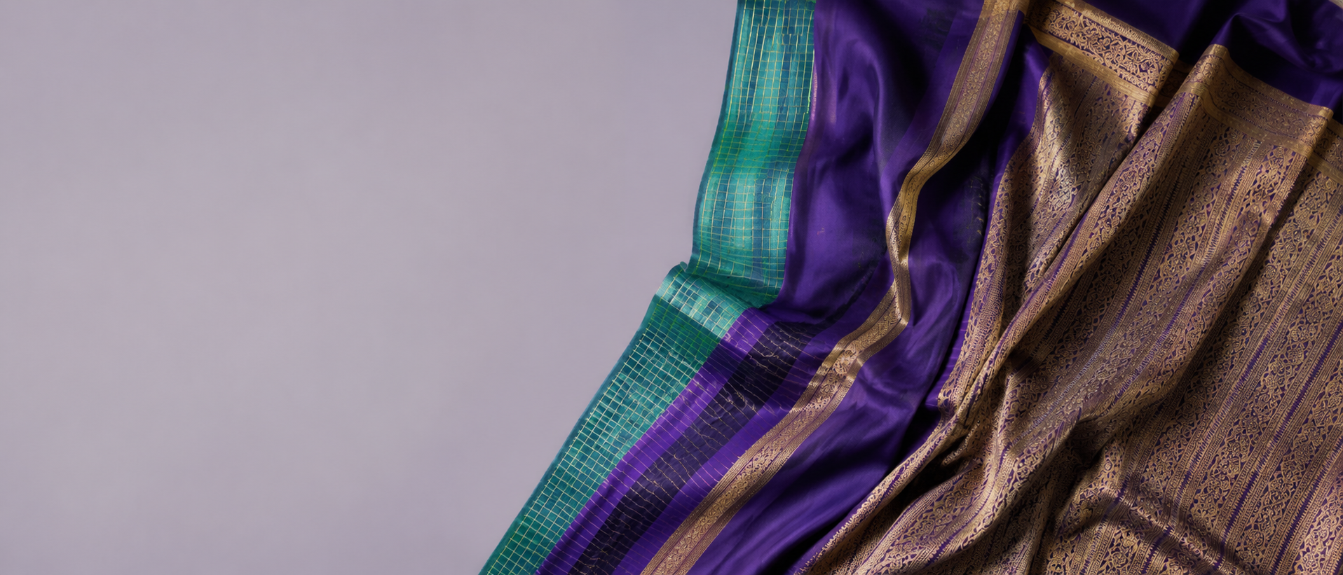Purple silk saree with a gold zari border and teal pallu edge, draped against a lavender backdrop